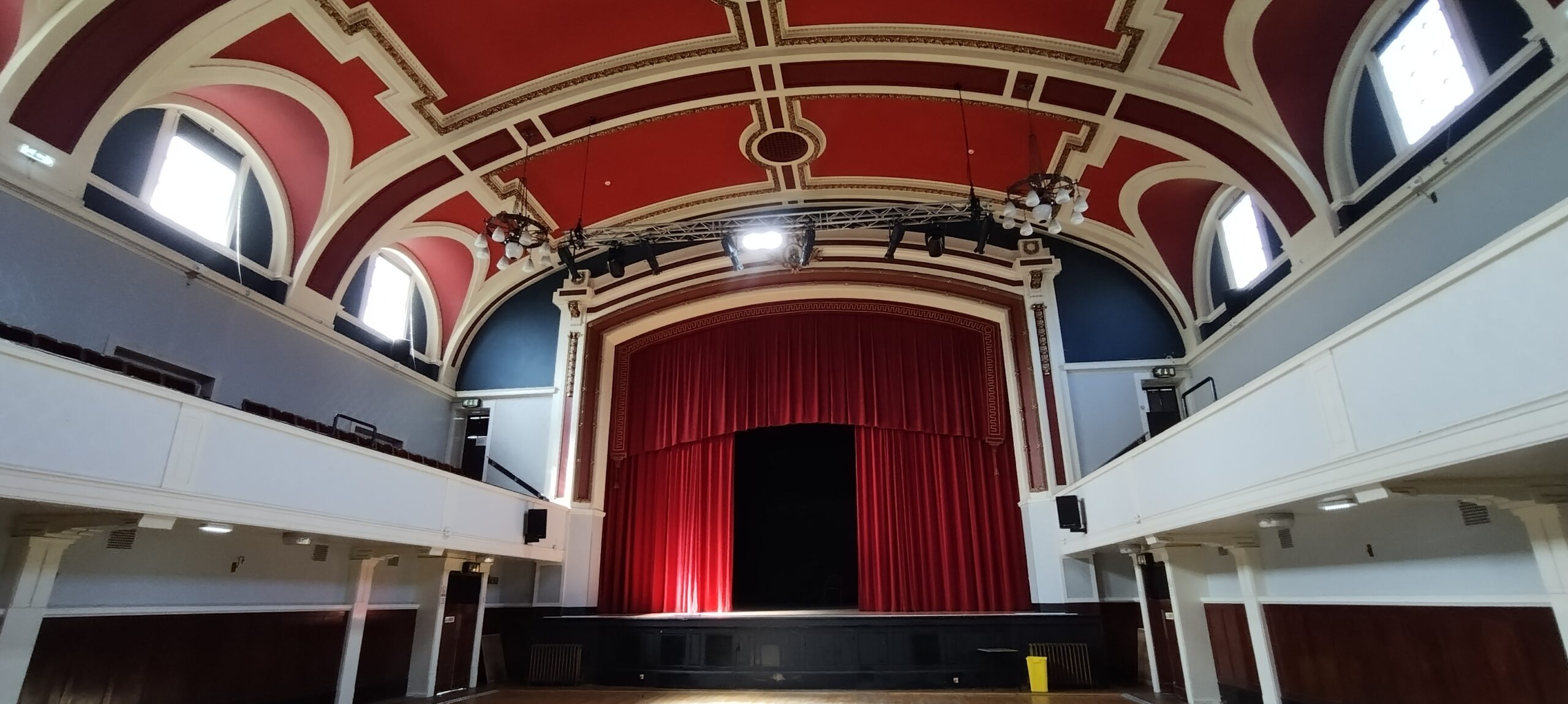 A photograph showing an auditorium. There is a stage with a proscenium arch, lined with red curtains.