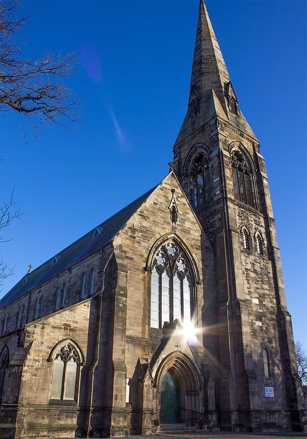 A photograph of a church with a 165ft spire, built from Binny Stone in the Early Decorated style. The sun is reflecting off the large stained glass window above the main doors.